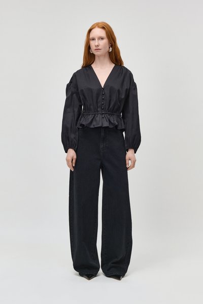 Femme aux longs cheveux roux portant un chemisier noir boutonné avec des manches bouffantes et un pantalon noir à jambes larges, debout devant un fond uni.