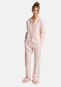 Lichtroze pyjamastel met verticale strepen, bestaande uit een knoop-top en lichte broek, gecombineerd met pluizige roze slippers.