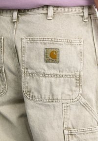 Beige Baumwoll-Shorts mit strukturierter Oberfläche, ausgestattet mit großen seitlichen Taschen und einem quadratischen Carhartt-Logo-Patch in Gelb und Beige.