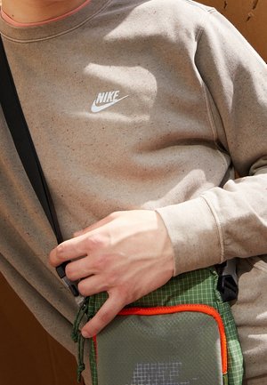 Personne portant un sweat-shirt Nike beige et tenant un sac bandoulière vert avec bordure orange et sangle noire.