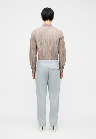 Άνδρας που φοράει ανοιχτόγκρι παντελόνι, μακρυμάνικο ριγέ πουκάμισο που έχει μπει μέσα στο παντελόνι και λευκά αθλητικά παπούτσια, στέκεται γυρισμένος με την πλάτη προς τα εμπρός σε λευκό φόντο.