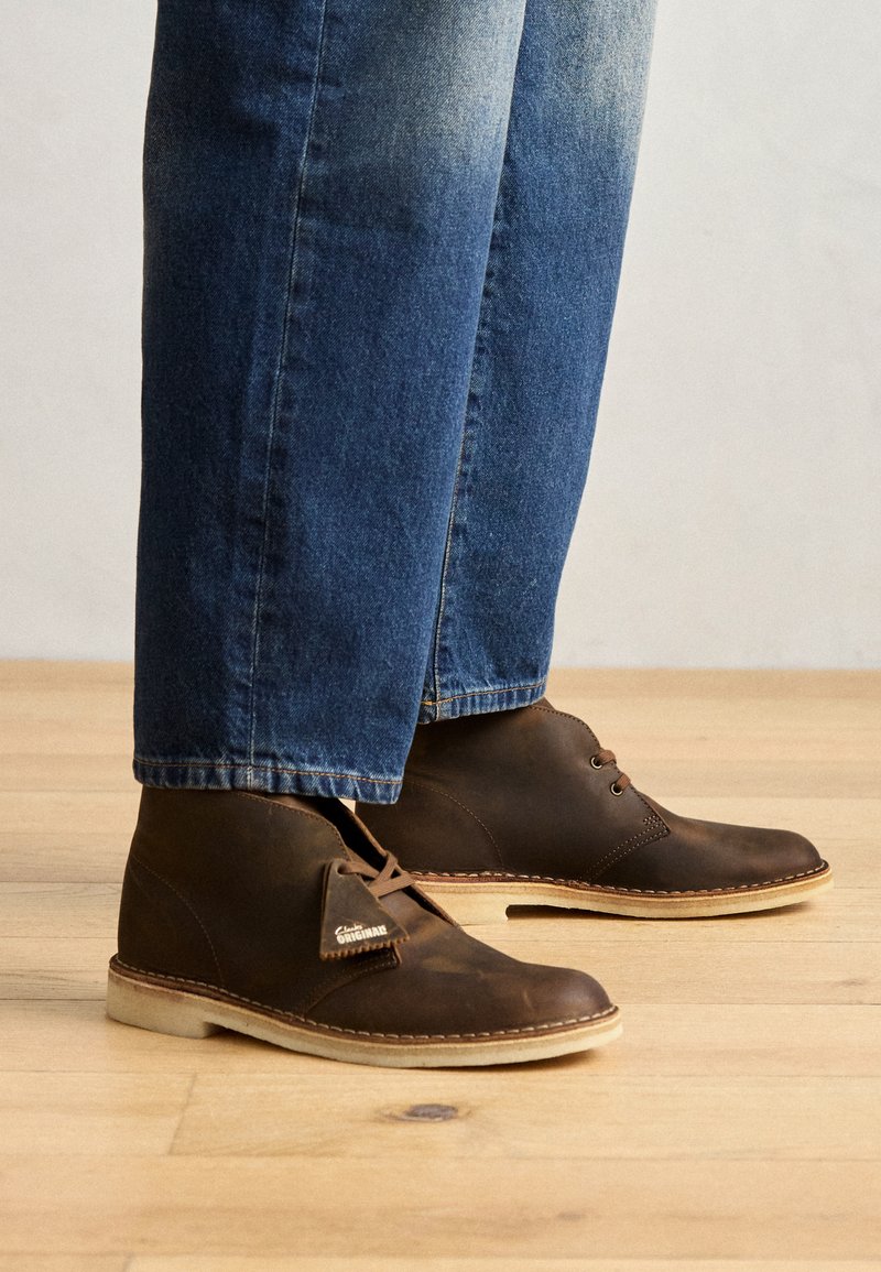 Stivali chukka in pelle marrone con punta rotonda, due occhielli per i lacci e suola in gomma naturale, abbinati a jeans di denim blu. Sfondo in legno.