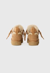 Pantofi din suede maro cu accente crem, căptușeală pufoasă și tălpi groase bej. Acestea au șireturi împletite și un logo UGG pe toc.