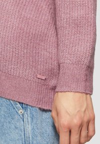 Gebreide trui in zachtroze met ribbelboorden en zoom. Heeft een klein logo-tag in dezelfde kleur nabij de zoom. Hand tegen denimstof.