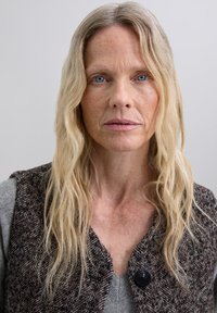 Persona de cabello largo que lleva un chaleco de espiga marrón sobre una prenda gris. El fondo es de un gris claro, centrándose en el rostro del sujeto.