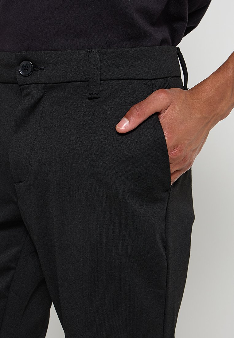 Schwarze Hosen mit klassischem Design, aus einem glatten Stoff gefertigt, mit einem Knopfdetail an der Taille und einer Hand in der Seitentasche.