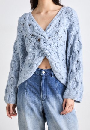 Femme portant un pull en maille épaisse bleu clair avec un devant torsadé sur un jean bleu délavé taille haute.