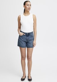 Haut blanc avec des détails en volants, short en denim bleu taille haute et ballerines noires. Le modèle se tient avec les mains dans les poches contre un fond neutre.