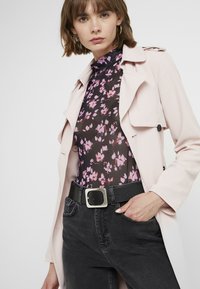 Trench rosa claro superpuesto sobre una blusa negra transparente con estampado floral y acentos rosas. Cinturón negro con hebilla cuadrada plateada, combinado con pantalones oscuros.