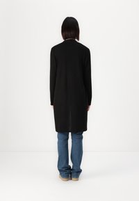 Black long cardigan with a straight silhouette, ribbed knit texture, and long sleeves, paired with blue denim jeans and beige shoes.