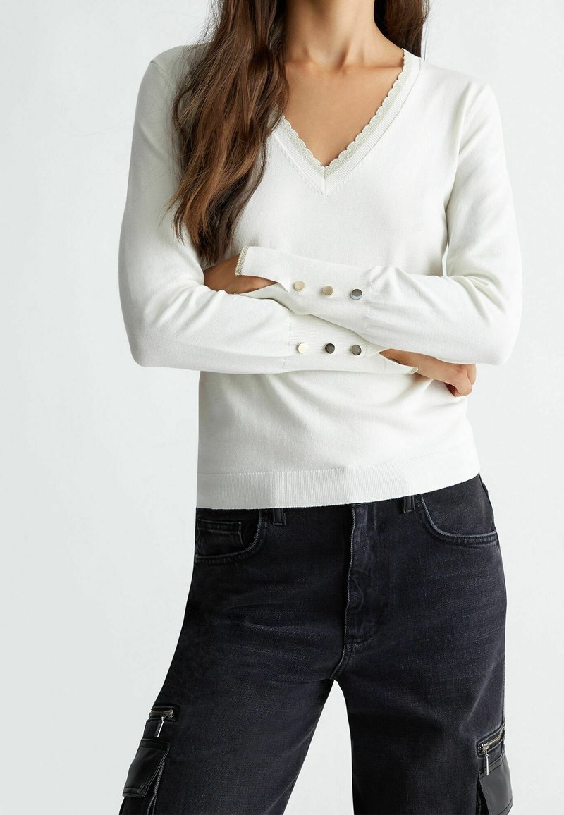 Bílý pletený svetr s výstřihem do V, vlnitým lemem a knoflíkovými detaily na rukávech. Nosený s tmavými kalhotami ve stylu cargo.