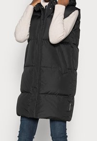 Gilet imbottito nero con cappuccio, caratterizzato da una chiusura con zip sul davanti, tasche laterali e una vestibilità ampia. Realizzato in un materiale liscio e trapuntato.
