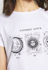 Vit bomull t-shirt med svarta grafiska tryck av himmelska motiv: halvmåne, sol med strålar och texten "COSMIC LOVE" ovanför "YOU," "ME," "THEM."