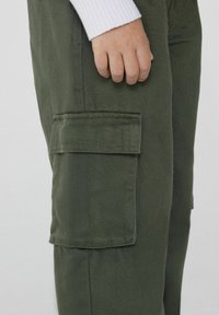 Olivově zelené cargo kalhoty s texturovanou látkou, které mají boční kapsu a přiléhavý střih. Z vrchu vykukuje bílý dlouhý rukáv.