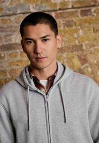 Grauer Reißverschluss-Hoodie mit Kordelzügen, über einem roten karierten Hemd getragen. Glatte Stofftextur, vor einer rustikalen Steinwand abgebildet.