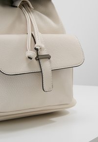 Mochila de cuero beige con cierre de solapa, parte superior con cordón y detalles de ribete negro. Textura suave y diseño aerodinámico.
