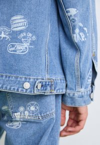 Light blue denim jacket featuring multiple white printed graphics and logos, snap buttons on cuffs, and a relaxed fit design.