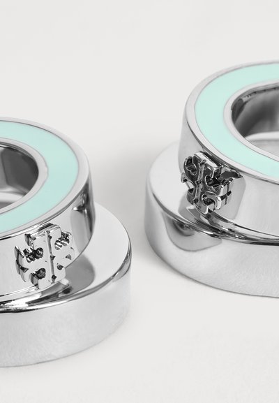 Deux bagues en métal argenté avec incrustation d’émail bleu clair et un emblème décoratif en forme de croix sur fond blanc.