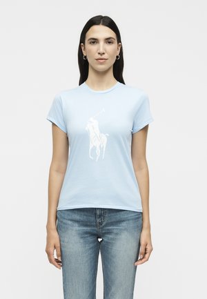 Γυναίκα με ίσια σκουρόχρωμα μαλλιά φοράει ανοιχτό μπλε μπλούζα με λευκό γραφικό παίκτη πόλο και μπλε τζιν, στέκεται μπροστά σε μονόχρωμο φόντο.