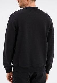 Sudadera negra con mangas largas, cuello y puños acanalados, y un tejido texturizado. La parte posterior presenta un diseño simple sin logotipos.