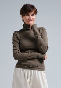 Brauner gerippter Stehkragenpullover aus elastischem Material, mit langen Ärmeln und tailliertem Design, kombiniert mit hellen, lockeren Hosen.