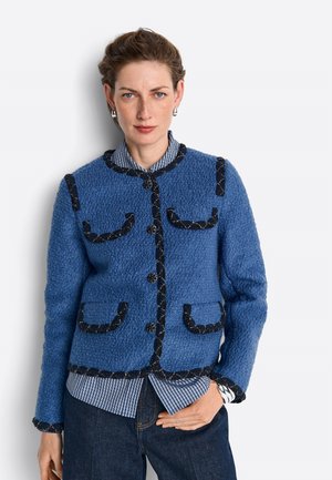 Donna che indossa una giacca blu strutturata con rifiniture nere sopra una camicia a righe e jeans scuri, in piedi con una mano in tasca.