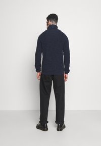 Homme de dos portant un pull col roulé bleu marine, un pantalon noir avec des ourlets et des chaussures noires sur un fond clair uni.