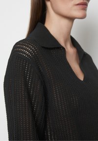 Schwarzer gestrickter Pullover mit einer Netzstruktur und einem kragenförmigen Ausschnitt, der einen V-förmigen Schnitt und strukturierte Schultern aufweist.