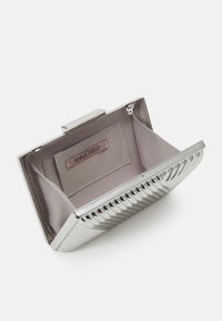 Kompaktowa metaliczna torebka kopertowa w kolorze srebrnym z żeberkowym wzorem, otwarta, aby ujawnić jasnobeżowe wnętrze z materiału oraz małą metkę z napisem "ANNA FIELD."