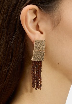 Zbliżenie ucha z złotymi, teksturowanymi kwadratowymi kolczykami z pięcioma wiszącymi niciami małych brązowych koralików.