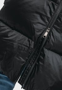 Schwarze Pufferjacke mit glatter, glänzender Textur. Sie verfügt über einen silbernen YKK-Reißverschluss und ein leicht geometrisches Muster auf dem Stoff.