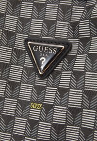 Musta kuvioitu kangaskassi, jossa on kolmiomainen musta ja kultainen "GUESS" -logo ja keltainen teksti sekä jäsentelty geometrinen muotoilu.