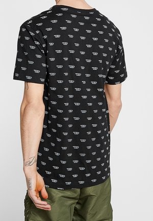 Personne portant un t-shirt noir avec un motif répété de logo blanc et un pantalon vert olive, vue de dos.