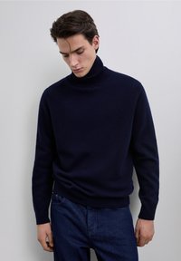 Pull à col roulé bleu marine en tissu tricoté doux, avec un col et des poignets côtelés, porté avec un jean en denim foncé.