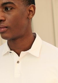 Camisa polo blanca con un cuello abotonado, con una textura suave y un sutil detalle de costura. Primer plano del escote y parte de la cara.
