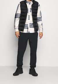 Gilet imbottito nero sopra una camicia bianca con motivi a quadri blu e grigi, abbinato a pantaloni neri e sneakers nere.