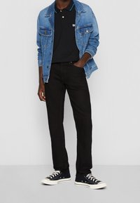 Chaqueta de mezclilla en azul claro, polo negro con cuello, jeans negros y zapatillas bajas negras con suelas blancas. Atuendo casual, diseño ajustado.