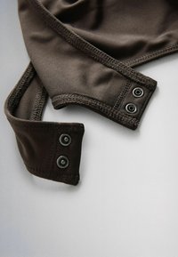 Primer plano de tela marrón oscura con bordes reforzados cosidos y cierres de botón a presión de metal en dos correas superpuestas.