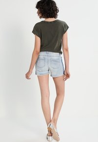 Olivově zelený top s krátkým rukávem, světle modré džíny šortky s třepenými okraji a stříbrné sandály. Jednoduchý, ležérní design s minimalistickými detaily.