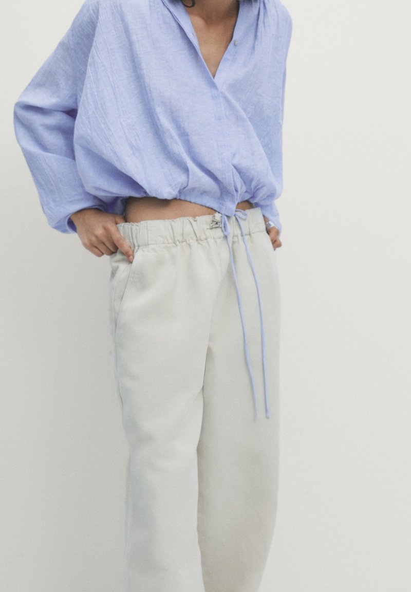 Haut croisé en lin bleu clair avec une coupe ample et un ourlet froncé. Pantalon crème avec cordon de serrage, poches latérales et silhouette décontractée.