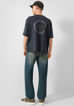 Man met een zwarte pet, zwarte grafische t-shirt met de tekst "Everyday Ambition", vervaagde blauwe spijkerbroek en bruine sneakers, staand met zijn rug naar de camera.