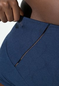 Ręka ściągająca pas ciemnoniebieskiej bezszwowej bielizny z siateczkowym panelem na ciemnej skórze tułowia.