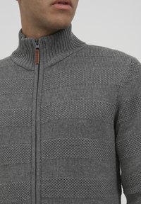 Grauer Reißverschluss-Pullover aus gestricktem Material mit geripptem Kragen. Verfügt über ein texturiertes Muster mit horizontalen Streifen und einem braunen Reißverschlussanhänger.