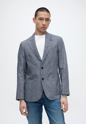 Mladý muž v sivom károvanom saku cez bielu košeľu a modré džínsy stojí pred jednofarebným bielym pozadím.