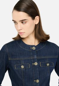 Džínová bunda v tmavě modré barvě s kulatým výstřihem, zlatými knoflíky, dvěma náprsními kapsami a kontrastními švy.