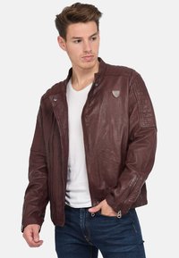 Junger Mann mit offenem braunem Lederjacke über einem weißen Hemd und blauen Jeans, der nach links schaut und die Hand in der Tasche hat.