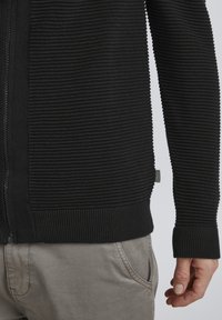 Schwarzer gerippter Pullover mit durchgehendem Reißverschluss, strukturiertem Stoff und schmalen Bündchen, kombiniert mit hellgrauen Hosen.