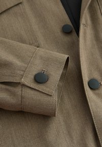 Brown fabric jacket with a textured finish, featuring black button accents on the cuff and a detailed seam along the sleeve.