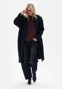Zwarte oversized jas over een donkerpaarse trui, gecombineerd met losse zwarte jeans en zwarte puntige laarzen. Gladde stof, minimaal ontwerp.