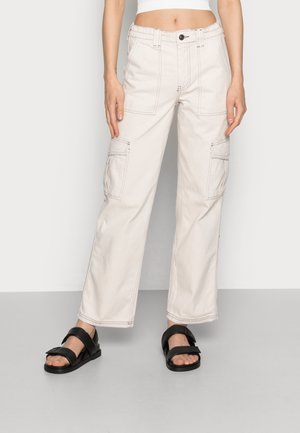 Kvinde iført off-white vidtbenede cargobukser med for- og sidelommer med klap og sorte åbne sandaler, stående på gråt gulv.